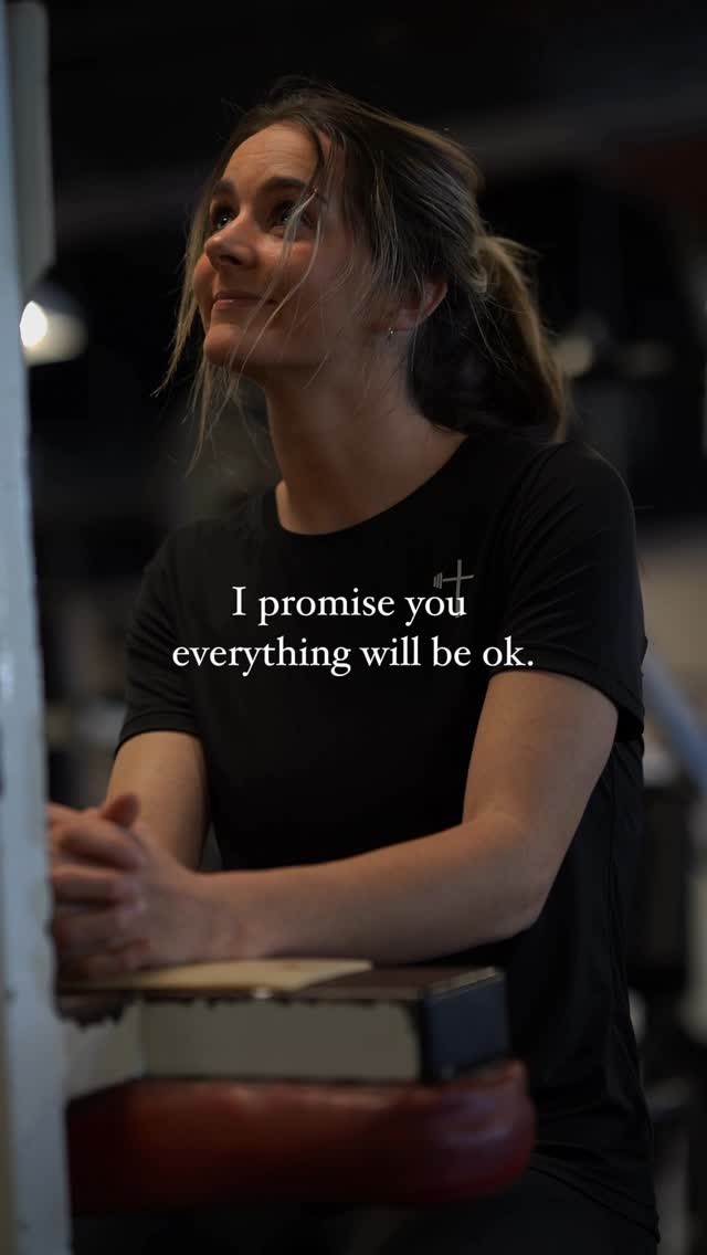 A woman with light brown hair pulled back in a partial ponytail is looking up and to her right with a slight smile, appearing contemplative. She's wearing a black t-shirt with a small white cross graphic. Overlayed on the image is white text on a dark background, reading: "I promise you everything will be ok." The overall lighting is soft, and the background is blurred.