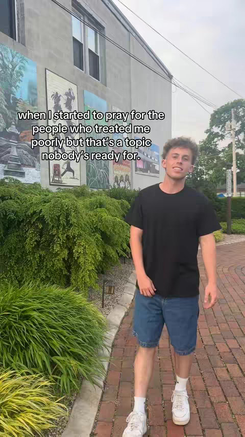 A young man in a black t-shirt and jean shorts stands on a brick walkway in front of lush green bushes and a building with murals. Text overlay reads: when I started to pray for the people who treated me poorly but that's a topic nobody's ready for.