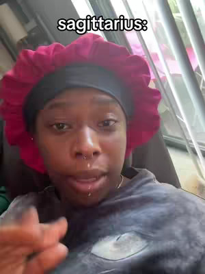 A close-up shot features a person with dark skin looking directly at the camera with a serious expression. The person is wearing a dark-colored head covering, and a red, ruffled headscarf. The background includes window and indoor environmental details, with a shallow depth of field.