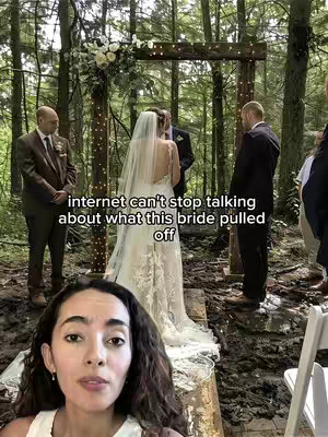 A selfie style video featuring a woman with dark hair in the bottom of the frame, looking at the camera. In the background is a wedding scene; a bride is in the center, with a man standing behind her, and two other men on either side of her. The scene takes place outdoors in a wooded area.