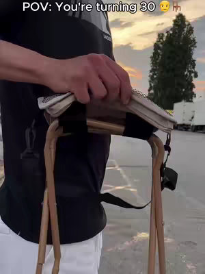 A person, seen from the torso down, is walking, holding a small wooden stool with a canvas seat. The background depicts a parking lot with a treeline and a partially visible sunset.