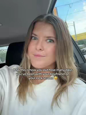 A woman with blonde hair and a white sweater looks towards the camera with a slightly concerned expression while sitting in a car. Text overlays inform the viewer how to display how many days they've been with their boyfriend on their lock screen.