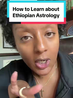 A selfie-style video cover featuring a woman with curly dark hair, looking directly at the camera and gesturing with her hand. Above her head in a turquoise rectangle is the text "How to Learn about Ethiopian Astrology". She has fair skin, and is wearing a black top along with a gold necklace with a cross pendant and a large gold ring on her finger. The background is out of focus, suggestive of a home setting, with a framed picture visible.