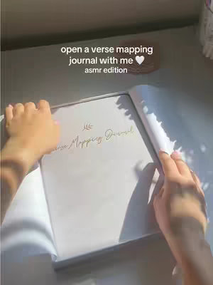 The cover image shows a person opening a verse mapping journal and their hands are visible. The hands are holding the book open from both sides, revealing a blank page and the title of the journal. The background is a light-colored surface with softer lighting.