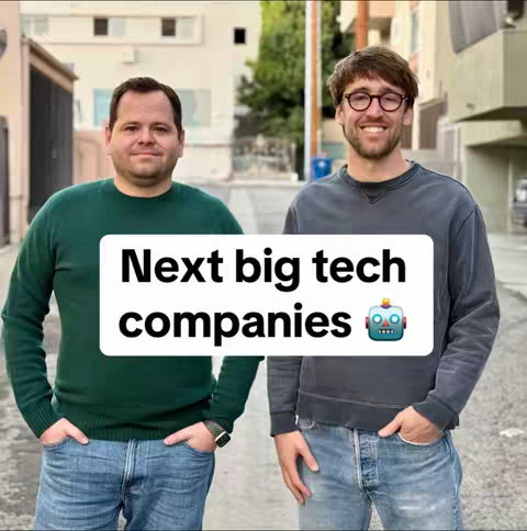 Two men are posing in what appears to be an alleyway of businesses. They are standing side-by-side, looking at the camera and smiling. The man on the left is wearing a green sweater, and the man on the right is in a grey sweatshirt. In front of them is a white and black box with the words, "Next big tech companies" and a robot emoji.