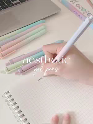 The image shows a person's hand writing with unique gradient gel pens on a white notebook. The pens are arranged on a white desk next to a silver laptop and other stationery. The overall aesthetic is clean and organized.