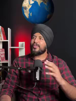 A man wearing a turban and a plaid shirt speaks into a microphone. Behind him is a globe decoration and a bookshelf, with the background in a dark red color. The lighting is dim, and the overall composition suggests a podcast or video recording setting.