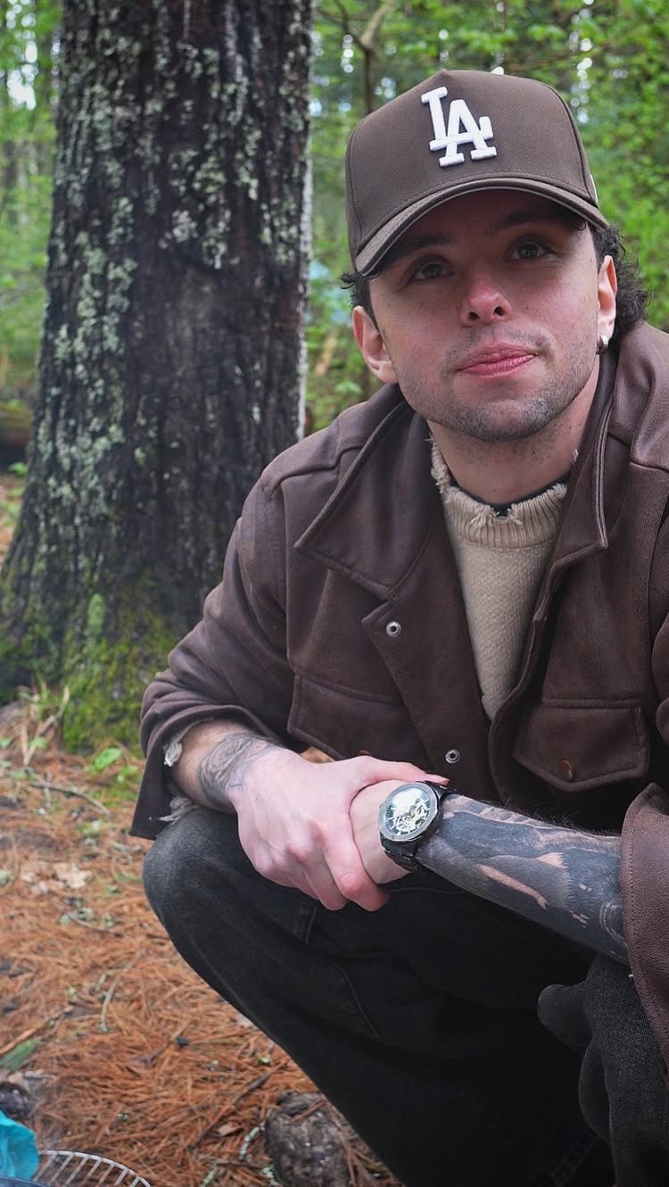 A man, wearing a brown LA Dodgers cap and a brown jacket, crouches near a tree in a forest setting. He has a watch on his wrist and is looking towards the camera.