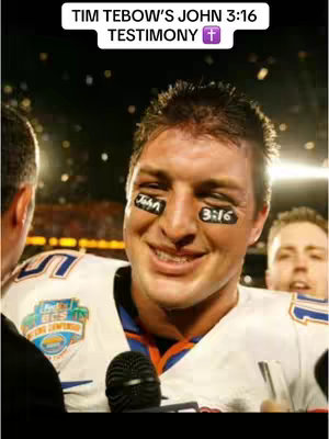 The cover image shows a man, presumably Tim Tebow, smiling and being interviewed. The man is wearing a white and blue football jersey. There are black marks under his eyes, with "John" written on one eye and "3:16" on the other. A microphone is close to his face. Another man is partially visible on the left side of the frame, and a person is behind the main person on the right. The background is blurred. Above his head is white text that reads "TIM TEBOW'S JOHN 3:16 TESTIMONY" with a purple cross.