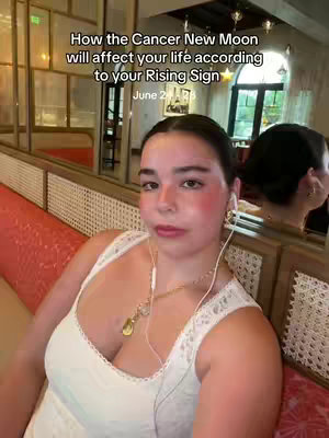 The cover image shows a woman with a makeup look, sitting on a red-cushioned bench indoors next to a window. A text overlay at the top states, "How the Cancer New Moon will affect your life according to your Rising Sign". The woman is wearing a white top and has a gold necklace. The background has a blurred interior design that includes the light and shadow effects.