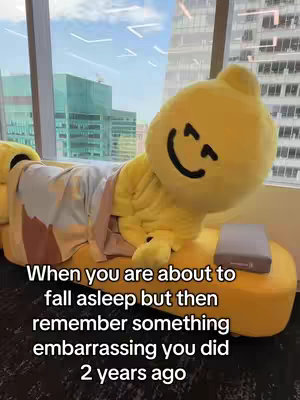 The image shows a yellow, blob-shaped character, possibly a mascot, reclining on a yellow couch by a large window. The character is wearing a robe and has a drawn-on face with a large smile, suggesting a sleepy or relaxed state. The background features a cityscape with tall buildings and a clear sky, giving the scene a bright and airy feel. Text overlays, placed beneath the character, address a relatable experience about recalling an embarrassing moment. The composition is simple, with the figure centered and the text clearly visible.