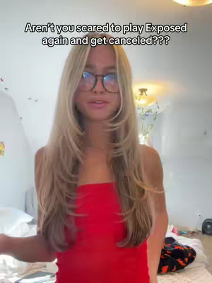 A young woman with long blonde hair and glasses stands in front of a messy white wall. She is wearing a red tube top. Text overlayed on the image reads "Aren't you scared to play Exposed again and get canceled???"