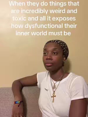 The cover image features a woman with dark skin and a patterned headscarf, sitting on a gray couch against a beige wall. She is wearing a white t-shirt and several necklaces, including one with a cross. She has a neutral expression and is looking slightly to the side. The overall composition is a medium shot, focusing on the woman's upper body and facial expression.