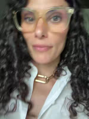 A close-up shot of a woman with curly hair and glasses looking at the camera. She's wearing a white shirt and a gold necklace. The background is blurred, suggesting an indoor setting.