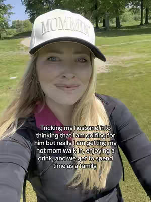 A woman wearing a white cap with the word "Mommin" on it, standing outside on a golf course. She is smiling faintly, and the background includes green grass and trees.