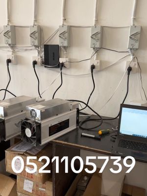 The image is a product shot featuring several cryptocurrency mining machines set up in a room. The machines are large, metallic gray, with fans visible. They are plugged into electrical outlets on a white wall, with multiple power cords and wires visible. A laptop is open on a table to the right of the machines.  Below, a phone number is superimposed in white. The overall setting looks like a technology or electrical installation space.