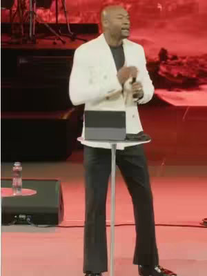 A man in a white blazer and black pants is speaking into a microphone on a stage. A lectern with a tablet sits in front of him. The background is blurred red.