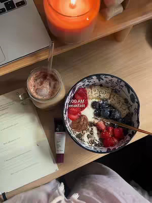 A top-down shot showing a workspace with food. A bowl of breakfast sits in the center, with berries and nuts. To the left is a glass of iced coffee, a laptop, and a tube of beauty cream. A lit candle is on the wooden surface in the background.