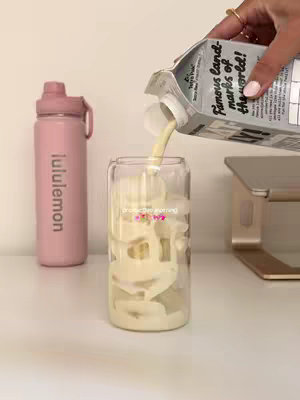 A person is pouring a white liquid, possibly milk or a smoothie, into a clear glass filled with what appears to be ice cubes. To the left is a pink water bottle, and on the right is a laptop stand. The background is a plain, light-colored surface, creating a clean and minimalist aesthetic.
