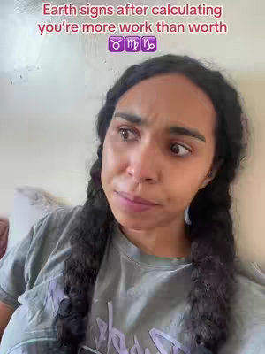 A selfie-style shot focuses on a woman's face, framed from the chest up. She has long, dark braided hair and is wearing a gray t-shirt. The image has text overlay at the top in pink, with astrological signs. The woman has a concerned or contemplative expression, with her gaze slightly averted.