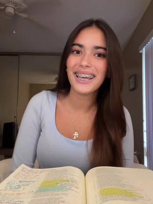 A young woman with long brown hair and braces is the central figure, seated and facing the camera with a gentle smile. She is wearing a light blue top and a necklace. In the foreground is an open book with highlighted text.