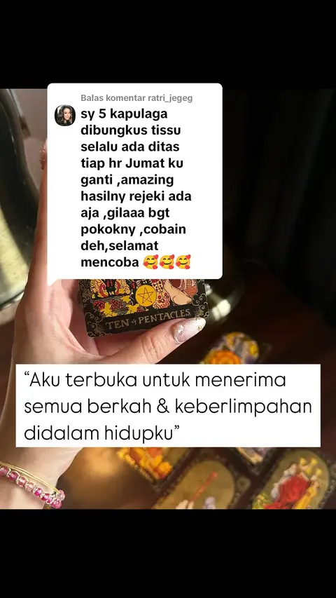 A hand holding a tarot card with the text "TEN of PENTACLES" and a comment from ratri_jegeg overlayed on the image in a rectangular white box. Below the card is a text in a white box that reads "Aku terbuka untuk menerima semua berkah & keberlimpahan didalam hidupku". The cover has a dark and blurry background.