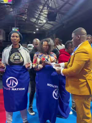 A group of people, likely in a church or event setting, are gathered together. Three individuals in the center are holding up blue fabric aprons with a logo. The background includes a blurred view of other people, and there is a lighting fixture overhead.