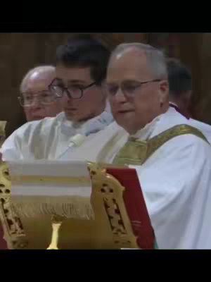 A medium shot shows several people in religious attire. A man in a white robe with a gold decorative collar is speaking and reading from a red book on a decorative stand. Behind him, a younger man in glasses and a white robe looks on, and a third man with glasses is partially visible in the background.