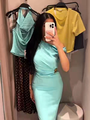 A woman is taking a selfie in a clothing store. She is holding up her phone with her face partially obscured. She is wearing a light blue, one-shoulder dress with a gold buckle. Behind her, there are clothing racks with hanging clothes in a changing room. The color palette is soft, and the lighting is from above. The composition presents a stylish outfit.