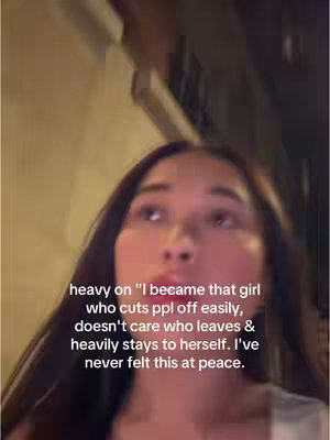 The image shows a young woman from a low angle, looking upwards with a slightly open mouth, set against a blurry, warmly lit background suggesting an outdoor setting. She has dark hair and is wearing a light-colored top. Text is overlaid, expressing sentiments of detachment and self-reliance.