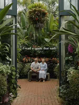 The image shows a couple sitting on a bench in a lush, green garden setting. Tall glass windows and doors flank a pathway leading into the scene, with abundant vegetation creating a natural archway. The couple appears relaxed and is looking up at the sky. The general composition is well-balanced, with natural light and a soft, muted color palette.