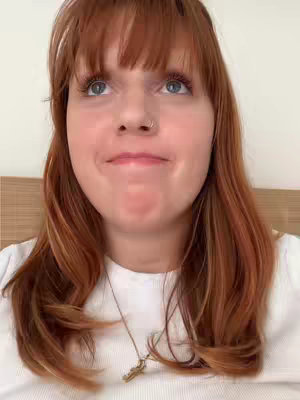 A close-up shot of a young woman with fair skin and red hair looking upwards with a slight smile. She's wearing a white shirt and a necklace with a small pendant. The background is a plain, light-colored surface.