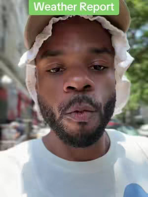 A medium shot features a Black man with a close-up view, focusing on his face in an outdoor setting. He is wearing a white t-shirt. He's wearing a brown hat with a white cloth around it. The background is slightly blurred, suggesting an unfocused street or outdoor environment. The man has a short, well-groomed beard and mustache. He has a neutral expression with his lips pursed. Above his head, a banner displays the text 'Weather Report' in a green rectangle.