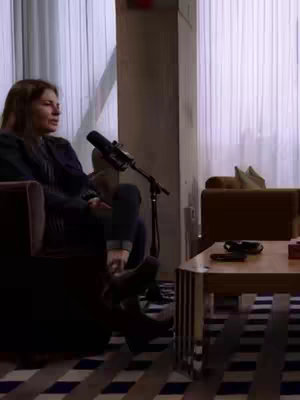 A woman sits on a couch in front of a closed window. A microphone is positioned in front of the woman. There is a small table to the right.