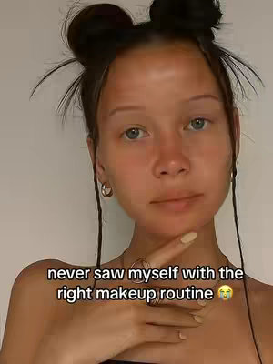 A selfie-style image of a fair-skinned person with blue eyes, their hair styled in two buns on top of their head. The person is holding their index finger up, and there is text overlaid on the image.