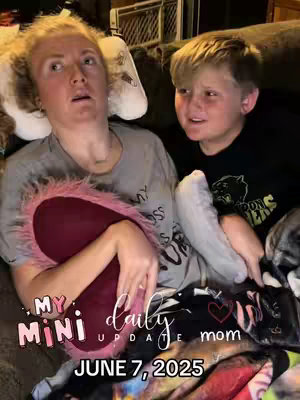 A woman and a young boy are seated close together on a gray sofa, likely indoors. The woman is looking upwards with a slightly concerned or emotional expression, her hands are holding a fluffy pink pillow. The boy is looking towards the left, seemingly watching something or someone off-camera and is wearing a black t-shirt. The foreground includes text overlays in a playful font: "MY MINI daily UPDATE mom," with "JUNE 7, 2025" below, and a small heart emblem.