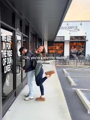A well-lit outdoor scene. The image features two people standing in front of a glass door. The person on the right is the main subject of the photo, and they are smiling and holding up two fingers. The background includes a winery sign, a building entrance, and a parking area.