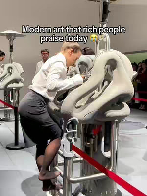 A woman is standing in front of sculptures inside an art exhibit. She is wearing a white shirt, black skirt, and pantyhose. The background includes additional spectators and the exhibit space itself. Text overlayed on the image reads 'Modern art that rich people praise today.'