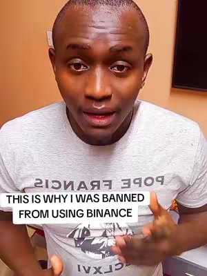 Close-up shot of a man in a light gray t-shirt, facing the camera. The text overlay at the bottom reads "THIS IS WHY I WAS BANNED FROM USING BINANCE". Background includes a beige wall and a glimpse of a dark-colored object on the right. The man appears to be speaking, possibly explaining the reason for his ban.