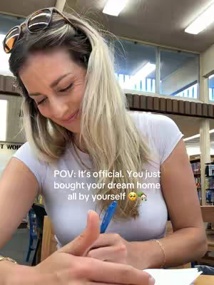 A woman with blonde hair and sunglasses on her head is seated indoors, writing on a piece of paper. She is smiling and appears content. Text overlays say "POV: It's official. You just bought your dream home all by yourself." 