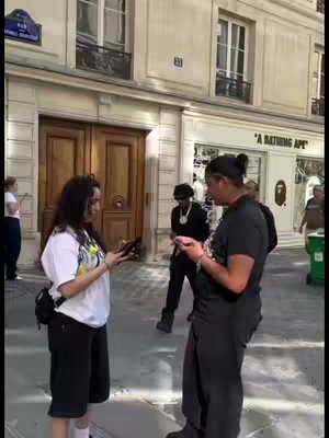 Two people are standing on a city street, each holding phones. They appear to be engaged in conversation beside a person wearing sunglasses and a black outfit in the near background. Behind them are buildings, a sign, and a store with an "A Bathing Ape" sign, suggesting a shopping or urban setting.