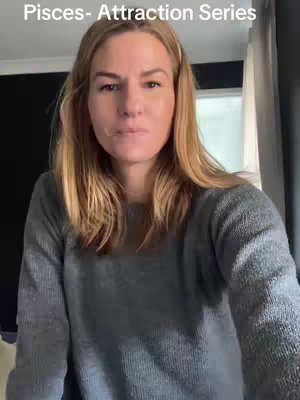 A medium shot featuring a fair-skinned woman with blonde hair looking directly at the camera. She has a neutral expression and is wearing a gray sweater. Background includes interior elements such as a window and curtains.
