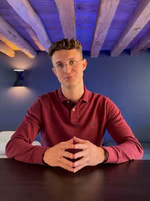 A man in a maroon polo shirt sits with his hands clasped in front of himself at a dark table. He wears glasses and looks directly at the camera. The background includes a blue wall and a wooden ceiling structure.