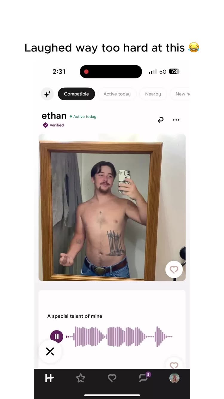 A screenshot from a dating app features a shirtless man taking a mirror selfie. The man is standing in a bathroom setting, with a wooden frame around the mirror. The app displays his profile, with a verification checkmark next to his name. The man has a tattoo of several vertical lines on his torso.
