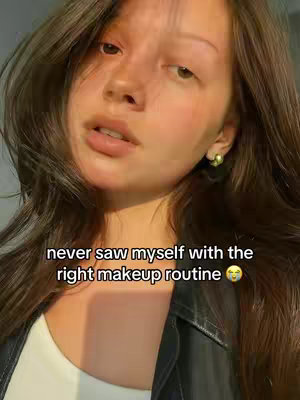A close-up selfie of a woman with long, wavy brown hair. She has smooth skin with subtle blush. The background is out of focus.