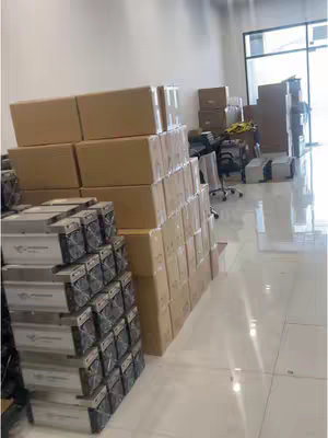 An indoor shot of a room with stacked boxes and cryptocurrency mining machines. The room is lit by bright overhead lights, and the floor is tiled in a reflective white. Stacks of brown cardboard boxes are visible throughout the room, alongside multiple rows of black and silver mining machines. The composition includes an open, expansive view of the space.