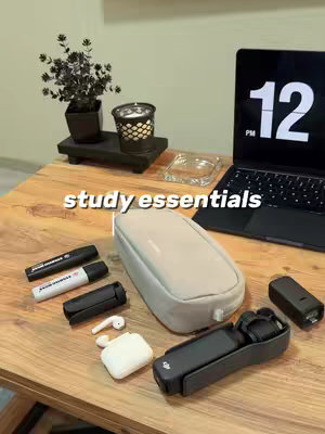A top-down shot of a wooden table displays a variety of study essentials. In the background is a laptop with a black screen displaying the time 12 PM. Several items are arranged on the table, including markers, a pencil case, a small black tripod, a white case with earbuds, a black case, and a small black item.