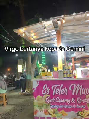An outdoor night scene with a food stall, the text overlay stating 'Virgo bertanya kepada Gemini'. The stall has a pink banner with text in Indonesian, and there are trees and lights in the background with some people.