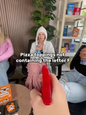 The image shows a person holding a red spray bottle in the foreground, seemingly pointing it at another person in the middle ground. The person in the middle ground is centered, wearing a white hoodie. There are two other persons in frame, and a background with shelves, plants and other items.