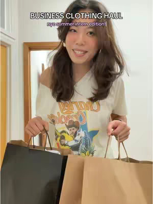 A young woman with long brown hair, wearing a white t-shirt and holding shopping bags. The background is a simple white wall. The text overlay reads 'BUSINESS CLOTHING HAUL nyc summer intern edition!'